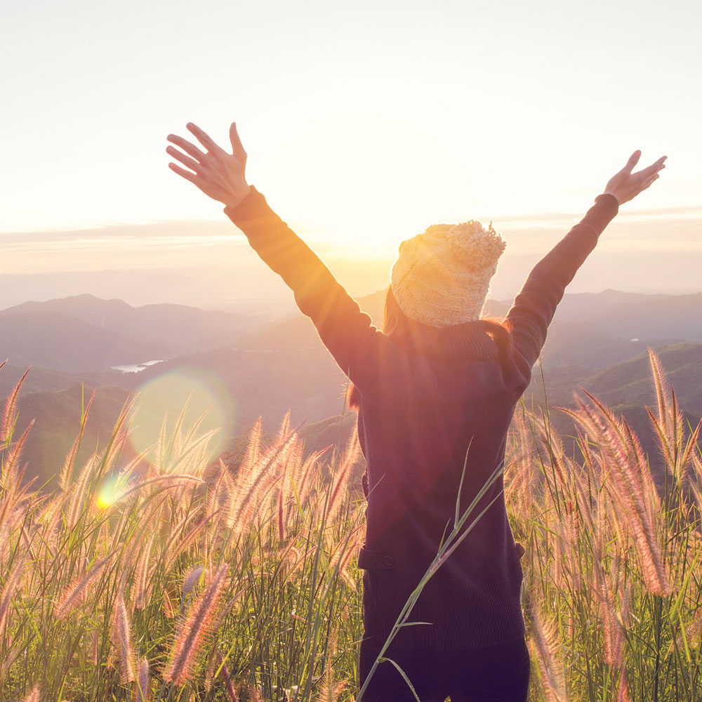 Carefree Happy Woman Enjoying Nature on grass meadow on top of mountain cliff with sunrise. Beauty Girl Outdoor. Freedom concept. Len flare effect. Sunbeams. Enjoyment.